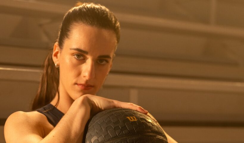Indiana Fever guard Caitlin Clark sits on bleachers holding a black and gold signature Wilson basketball.